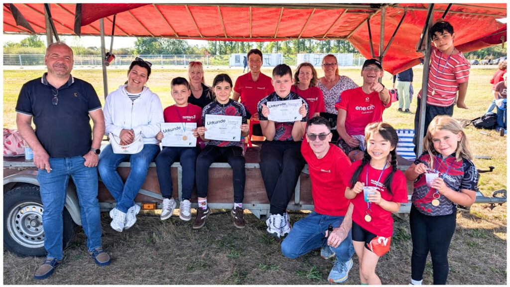 Gruppenbild mit Eltern, Trainern und Kindern nach dem Rennen. Alle blicken lachend in die Kamera. Kinder halten halten Urkunden und Medaillen hoch.